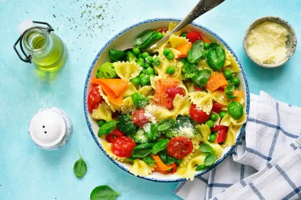 Un plato de pasta con verduras frescas: brócoli, guisantes, tomates cherry y zanahoria, espolvoreado con queso rallado.