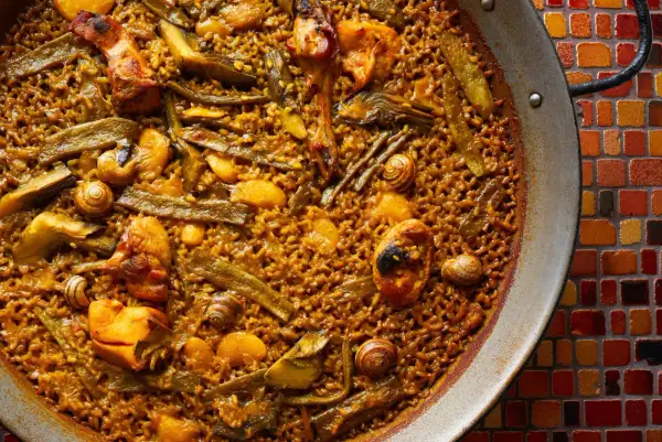Paella de pollo con trozos de pollo dorado, judías verdes y caracoles, servida en una paellera sobre un mosaico de colores.