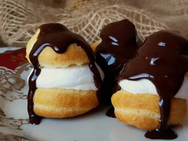 Deliciosos profiteroles de nata cubiertos con chocolate negro derretido, servidos en un plato blanco con detalles dorados.