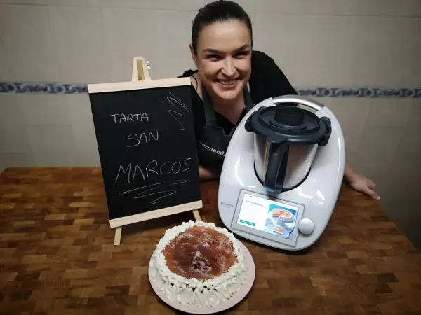 Mujer sonriente junto a una tarta San Marcos y una Thermomix. La pizarra anuncia "Tarta San Marcos".
