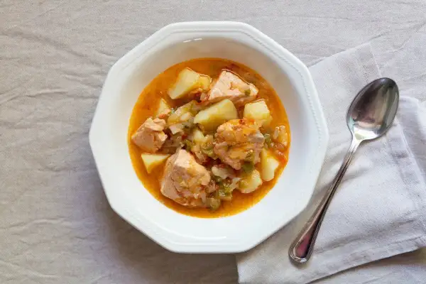 Un plato de marmitako de salmón con patatas y pimientos en una cazuela blanca, acompañado de una cuchara.