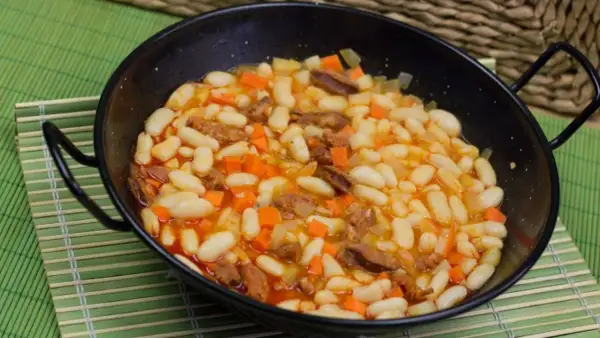 Un plato humeante de alubias con chorizo, zanahoria y patata en una paellera negra, servido sobre una esterilla verde.
