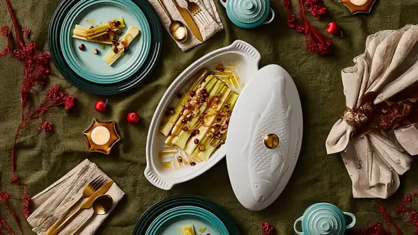 Puerros al horno, tiernos y dorados, servidos en una elegante fuente con tapa en forma de pez. Decoración festiva con velas y frutos rojos.