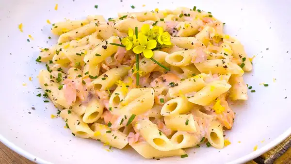 Pasta penne cremosa con trozos de salmón rosado, ralladura de limón y flores amarillas. Un plato delicioso.