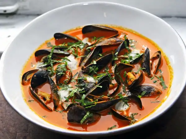 Un tazón blanco rebosante de mejillones al vapor en una salsa roja vibrante, adornado con hierbas frescas picadas.